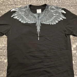 Svart t-shirt Marcelo Burlon M - Svart t-shirt från Marcelo Burlon County of Milan i storlek M. Snyggt tryck med gråa vingar runt halsen som ger en unik vibe. Tillverkad i mjuk bomull med klassisk passform och korta ärmar. Perfekt för dig som gillar streetwear med edge.