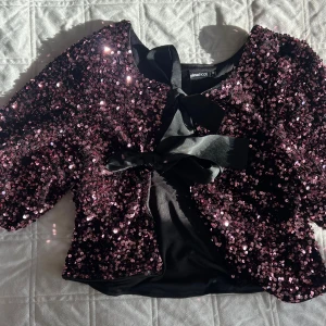 Glittrig paljettväst från Gina Tricot - Säljer en glittrig svart väst från Gina Tricot i storlek XS, täckt med mörklila paljetter och med en snygg rosett framtill. Perfekt för att sticka ut och ge din outfit extra edge. Västen har korta ärmar och är croppad i modellen.