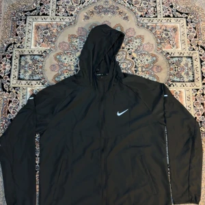 Svart Nike vindjacka med huva - Svart vindjacka från Nike med huva och dragkedja framtill. Jackan har reflekterande detaljer på ärmarna och klassisk Nike-logga på bröstet. Tillverkad i lätt polyester, perfekt för löpning eller utomhusaktiviteter. Snygg och sportig design.