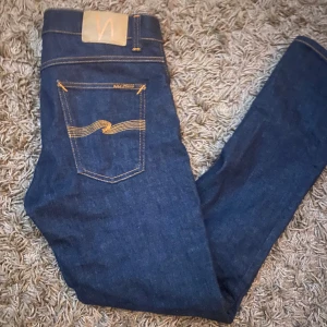 Mörkblå jeans från Nudie Jeans - Säljer ett par mörkblå jeans från Nudie Jeans med klassisk gul söm på bakfickan och läderpatch i midjan. Jeansen har en slim passform och raka ben, tillverkade i mjukt denimtyg som sitter snyggt och bekvämt.