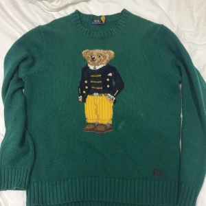 Grön stickad tröja Polo Bear Ralph Lauren - Grön stickad tröja från Polo Ralph Lauren med ikoniska Polo Bear-motivet framtill. Tröjan har rund hals, ribbade muddar och RL-broderi