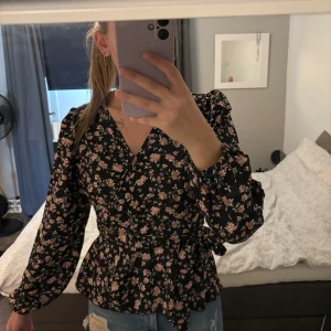 Blus - Säljer en svart blommig omlottblus med rosa och beige blommor. Blusen har v-ringning, långa puffärmar med resår vid ärmslut och ett snyggt knyt i midjan. Materialet känns lätt och flowy, perfekt till jeans eller kjol. Fräscht och trendigt mönster!
