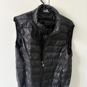 Svart dunväst från EA7 Emporio Armani - Snygg svart dunväst från EA7 Emporio Armani med quiltad design och hög krage. Västen har dragkedja framtill och EA7-logga på bröstet. Perfekt för lager-på-lager och ger en sportig vibe till din outfit.