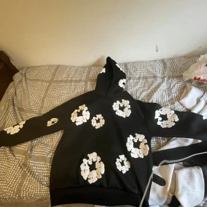 Svart hoodie Denim Tears med vita blommor - Svart hoodie från Denim Tears med stora vita blommor tryckta över hela tröjan, inklusive huvan och ärmarna. Klassisk känguruficka framtill och skön passform. Perfekt statement-plagg för dig som vill sticka ut.