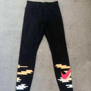 Svarta Nike träningsbyxor med tryck - Svarta leggings från Nike med färgglada grafiska detaljer i beige, vitt och rosa på benen samt en stor rosa Nike-logga. Tight passform och elastiskt material som passar perfekt till träning eller chill. Snygg och sportig look med hög midja.