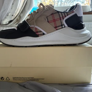 Burberry rutig sneakers med mockadetaljer - Säljer ett par snygga Burberry sneakers med klassiskt rutig mönster i beige, svart, vitt och rött. Skorna har mockadetaljer, snörning och en svart rem med Burberry-logga över foten. Platt vit sula och rund tå ger en modern vibe. Perfekta för dig som gillar statement-skor.