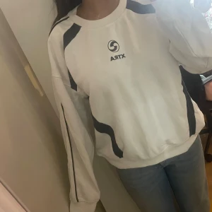 Vit sweatshirt från XTRA med svarta detaljer - Vit sweatshirt från XTRA med svarta grafiska detaljer på axlar och längs ärmarna. Tröjan har rund halsringning och långa ärmar med muddar. Perfekt för en sportig och avslappnad stil.