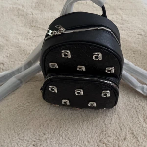 Svart ryggsäck från Alexander Wang - Cool svart ryggsäck från Alexander Wang i skinn med quiltad framsida och glittriga detaljer i form av små bokstäver. Väskan har två fack med silvriga dragkedjor och logga på sidan. Perfekt för dig som vill sticka ut med en trendig accessoar.