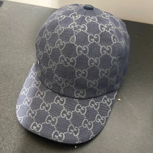 Gucci keps - Säljer en blå keps från Gucci med ikoniskt GG-mönster över hela kepsen. Kepsen är tillverkad i bomull och har justerbar rem baktill. Snygg och stilren accessoar för dig som vill sticka ut med lyxig streetstyle. 