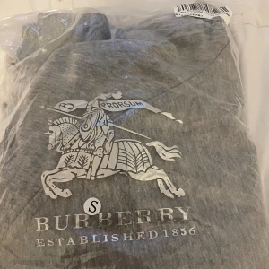 Grå hoodie med dragkedja från Burberry - Grå hoodie från Burberry med klassisk logga framtill, dragkedja och snörning i huvan. Insidan av huvan har det ikoniska rutmönstrets  Tröjan är i mjuk bomull och har långa ärmar samt fickor framtill. Perfekt för en chill och stilren look.