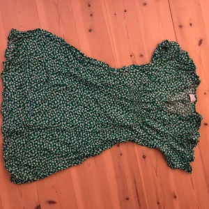 Grön blommig kortklänning Holly & Whyte - Säljer en grön kortklänning från Holly & Whyte by Lindex i storlek S. Klänningen har ett sött blommigt mönster i vitt och blått, v-ringad hals och korta ärmar med lätt volang. Materialet är mjukt och känns som bomull, perfekt för sommarens alla dagar.