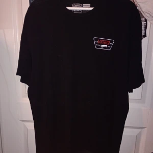 Svart Vans t-shirt i bomull XL - Svart klassisk t-shirt från Vans i storlek XL. T-shirten har korta ärmar och är tillverkad i 100% bomull. På bröstet finns en liten Vans-logga i vitt och rött. Perfekt för dig som gillar streetwear och enkel stil.