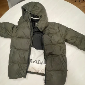 Olivgrön pufferjacka Calvin Klein Jeans - Säljer en olivgrön pufferjacka från Calvin Klein Jeans med huva och dragkedja. Jackan har stora quiltade partier, CK-logga på bröstet och ärmen samt svart innerfoder. Perfekt 
