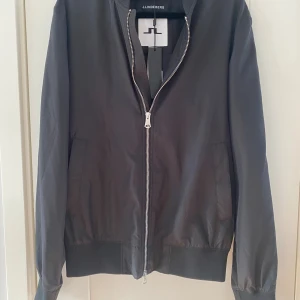 Svart bomberjacka från J.Lindeberg - Stilren svart bomberjacka från J.Lindeberg med klassisk ribbad krage, muddar och nederkant. Jackan har dragkedja framtill och två sidofickor. Materialet är lätt och känns skönt mot kroppen, perfekt för dig som gillar en clean look. Mått Över bröstet: ca 52 cm Längd från axeln och ner: ca 66 cm  Kommer från ett djur och rökfritt hem.