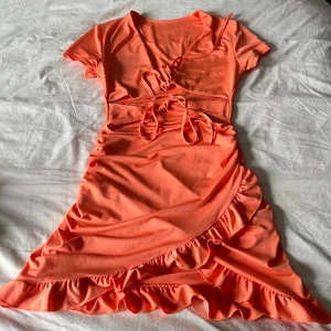 Kort orange omlottklänning med volang - Snygg kort klänning i stark orange färg med omlott-design och knytband i midjan. Klänningen har korta ärmar, volangdetaljer längs nederkanten och en cool cut-out vid midjan. Perfekt för dig som gillar att sticka ut med färg och detaljer.