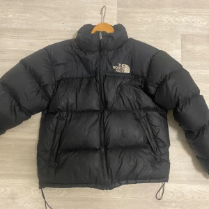 The North Face Nuptse 1996 jacka (M) - Säljer en svart dunjacka från The North Face i storlek M. Jackan är riktigt puffig med hög krage, dragkedja framtill och två snedställda fickor. Klassisk logga på bröstet och justerbar dragsko nedtill. Perfekt för kalla dagar. Storlek M ☺️🍂🍃