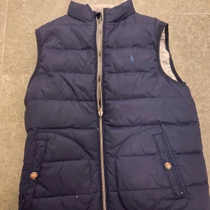 Mörkblå dunväst från Polo Ralph Lauren - Snygg mörkblå dunväst från Polo Ralph Lauren med quiltad design och klassisk ståkrage. Västen har två fickor med knappstängning och dragkedja framtill. Diskret Polo-logga broderad på bröstet. Perfekt att slänga över en hoodie eller t-shirt för extra värme. Model :163cm.   Barn jacka 