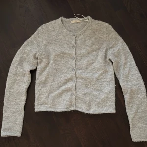 Grå kofta från Gina Tricot XS - Mysig grå kofta från Gina Tricot i storlek XS. Koftan har rund hals, långa ärmar och knappar framtill. Materialet är en blandning av ull, polyester, alpacka och elastan som ger en fluffig känsla. Perfekt för lager-på-lager under kyliga dagar. 