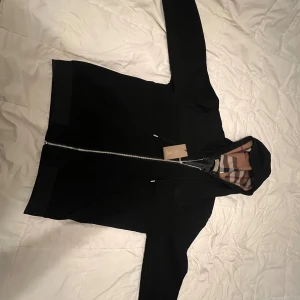 Svart Burberry hoodie med dragkedja - Svart hoodie från Burberry med klassiskt rutigt foder i huvan och insidan. Tröjan har hel dragkedja framtill, snörning vid huvan och ribbade muddar. Märkeslapp i nacken och snygga detaljer som ger en lyxig känsla. Perfekt för dig som gillar stilrena streetwear-plagg.