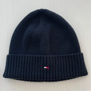 Mörkblå ribbstickad mössa Tommy Hilfiger - Snygg mörkblå ribbstickad mössa från Tommy Hilfiger med klassisk uppvikt kant och liten broderad logga framtill. Perfekt accessoar för kyliga dagar och passar dig som gillar stilren och enkel design.