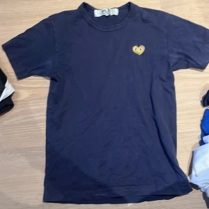 Mörkblå Comme des Garçons t-shirt - Snygg mörkblå t-shirt från Comme des Garçons med det ikoniska hjärtat i gult broderat på bröstet. Klassisk passform och rund halsringning, perfekt för en clean och stilren look.
