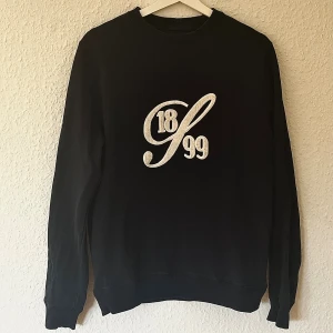 Mörkblå sweatshirt från Stenströms - Mörkblå sweatshirt från Stenströms med broderad text '1899' i vitt framtill och orange logga i nacken. Klassisk rund halsringning och långa ärmar. Perfekt för dig som gillar stilrena och tidlösa plagg. Bilden får tröjan att se svart ut, men den är marinblå/mörkblå.