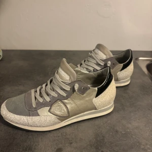 Philippe Model sneakers i grått och silver - Snygga Philippe Model sneakers i grått, silver och vitt med svarta detaljer på hälen. Skorna har en mix av skinn, mocka och textil, samt coola glittriga partier och klassisk snörning. Perfekta för dig som gillar en sportig och trendig look.