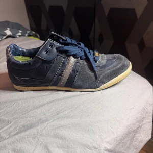 Blå sneakers från Hugo Boss - Snygga blå sneakers från Hugo Boss med detaljer i mocka och textil. Skorna har mörkblå snörning, gråa och blå paneler samt en beige sula. Sidan pryds av Hugo Boss-logga i silver och insidan har ett rutigt mönster. Perfekta för dig som gillar stilrena och sportiga skor.