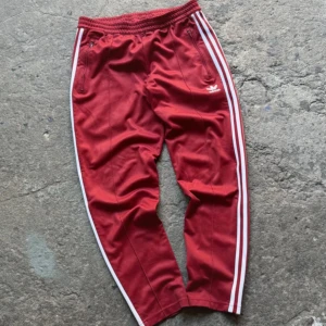 Röda Adidas trackpants  - Röda Adidas trackpants med klassiska vita ränder längs sidorna och broderad logga på låret. Dragkedjeförsedda fickor fram och en bakficka. Tillverkade i polyester för en sportig vibe.