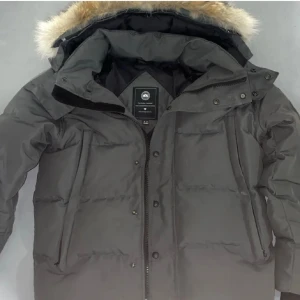 grå dunjacka från Canada Goose - Säljer en grå dunjacka från Canada Goose med stor huva och äkta pälsdetalj. Jackan har dragkedja, knappar och två stora fickor framtill. Klassisk patch på ärmen och quiltad design som håller dig varm hela vintern.