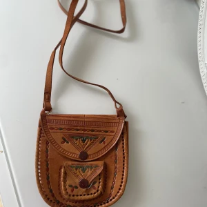 Brun axelväska i skinn med broderi - Unik brun axelväska i skinn med handgjorda broderade detaljer i grönt, rött och gult. Väskan har ett lock med tryckknapp, en mindre ficka framtill och dekorativa sömmar runt kanterna. Snyggt mönstrad och perfekt för dig som gillar vintagevibbar.