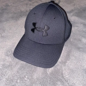Svart keps från Under Armour - Snygg svart keps från Under Armour med broderad logga framtill och diskret text baktill. Kepsen har böjd skärm och ventilerande paneler. Tillverkad i polyester för en sportig och bekväm känsla. Perfekt för dig som gillar stilren och enkel design.