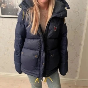 Mörkblå dunjacka från Fjällräven - Säljer en mörkblå dunjacka från Fjällräven med gul snörning och stor huva. Jackan har två stora fickor med knappar, dragkedja och Fjällräven-logga på bröstet. Perfekt för kalla dagar och riktigt snygg till jeans. Kan gå ner i pris för snabb affär 