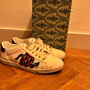 Gucci Ace sneakers  - Gucci Ace sneakers i vitt skinn med ikonisk broderad röd och svart orm på sidan. Baksidan har metallicröd och metallicgrön detalj med guldfärgad Gucci-logga. Klassisk låg modell med snörning och platt sula. Perfekta för dig som vill sticka ut med lyxig streetstyle.