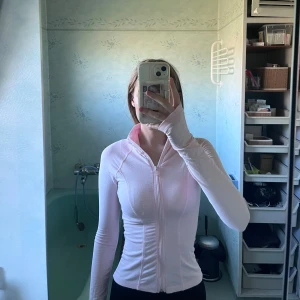 Ljusrosa zip-tröja med lång ärm - Säljer en ljusrosa långärmad zip-tröja med tight passform. Tröjan har dragkedja hela vägen framtill och tumhål vid ärmsluten. Perfekt för träning eller som ett snyggt lager till vardags. Enkel och stilren design.