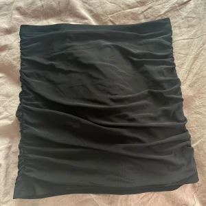 Svart kortkjol med rynkade sidor - Snygg svart kortkjol med rynkade sidor som ger en cool och trendig look. Kjolen har en tight passform och är gjord i ett mjukt syntetmaterial som sitter skönt mot kroppen. Perfekt att matcha med en crop top eller oversized hoodie.