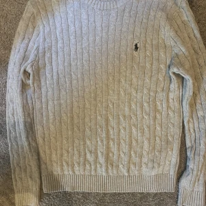 Ljusgrå kabelstickad tröja Polo Ralph Lauren - Kabelstickad ljusgrå tröja från Polo Ralph Lauren med klassisk rund halsringning och broderad logga på bröstet. Tröjan är i bomull och har ribbade muddar vid ärmslut och nederkant. Perfekt för lager-på-lager och snygg till jeans.