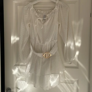 Vit kortklänning med volanger och bälte - Superfin vit kortklänning från NA-KD med volangkjol, långärmade transparenta ärmar och diskret prickigt mönster. Klänningen har resår i midjan, snyggt bälte med guldfärgat spänne och knytning vid halsen. Perfekt för sommarens alla tillfällen!