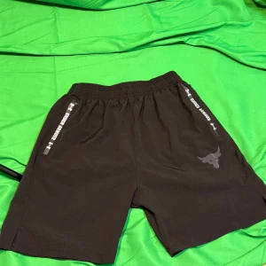 Svarta Project Rock short X under armour  - Svarta shorts från Project Rock med elastisk midja och snörning. Snyggt lila tjurhuvud-tryck på ena benet och dragkedjor med textdetaljer vid fickorna. Perfekta för träning eller chill. Materialet känns som polyester och passformen är normal.Liknande modell som originalet!