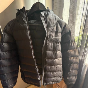 Svart dunjacka Peak Performance - Säljer en svart dunjacka från Peak Performance med huva och dragkedja. Jackan är quiltad och har en snygg, sportig look med diskret logga på bröstet. Perfekt för kalla dagar och enkel att matcha med allt.