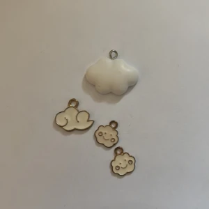 Vita molnberlocker i olika former - Fyra söta molnberlocker i vitt, perfekta till halsband eller armband. En är 3D-formad och de andra tre är platta med guldfärgade kanter, varav två har glada ansikten. Supergulliga detaljer för att skapa egna smycken.