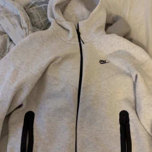Nike tech Vit - Nike hoodiejacka i ljusgrått med svarta detaljer och dragkedjor. Jackan har huva, hel dragkedja framtill och två fickor med svarta dragkedjor. Perfekt för en sportig och avslappnad stil.