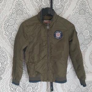 Sommarjacka  - En barn jacka perfekt för sommaren men bomber stil. Den har en lagad skada på insidan och en liten skada på utsidan men ej märkbar. Olivgrön utsida  Blå insida  Storlek 152  