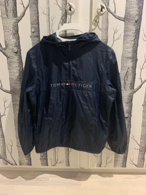 Tommy Hilfiger jacka  - Marinblå, tunn vindjacka i anorakmodell där man tar på sig den över huvudet.  Huva, dragkedja halvvägs och fickor. Tunna muddar i ärmslut och nedtill. Mycket fint skick! 