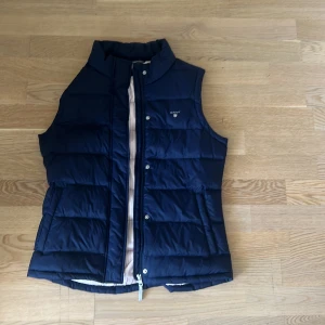 Gant väst  - En mörkblå väst från gant. Helt oanvänt alltså i nytt skick. Passar tonåringar är i storlek 176 passar även för de med storlek xs/s. Vita och rosa detaljer inne i jackan. Fickor utanpå samt i. 