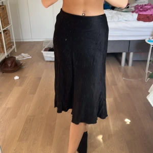 Svart midikjol  - Svart midikjol i satin från Zara i strl S. Har ett innerrum vilket gör den en aning tjockare. Stängs i vänstersida med en dragkedja. Lagat litet hål precis under dragkedjan, se bild 