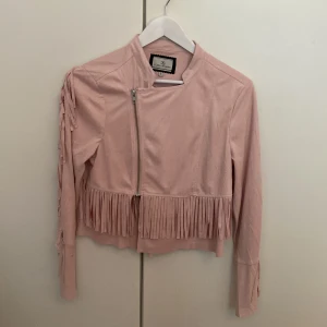 Jacka i mockaimitation med fransar - Blekrosa tunn jacka från Flash Woman i mockaimitation (polyester) med fransdetaljer. Storlek S/36. Fint skick.