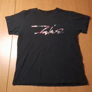 T-shirt - En svart t-shirt med tryck. I ganska bra skick men texten på framsidan har flagnat bort på vissa ställen. Den är 100% bomull.