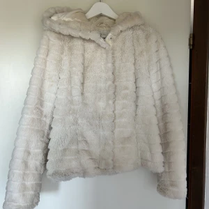 Lurrvig jacka  - En lurrvig vit midjelång jacka med mysig luva. Från Vila i storlek 38. Jackan är använd ett tag och har därför sminkfäckar på insidan av tyget. Det går nog bort i tvätten. Jag har inte tvättat pga av lurrvet. Men det ska nog gå bra! Jackan har inga fickor
