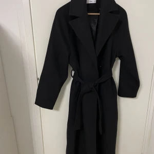 Ellos kappa Amina - Knappt använd, mycket bra skick Helt slutsåld, ord pris 1200 kr  Svart kappa i ullblandning. Stängning med två dolda tryckknappar. Knytskärp i midjan och två fickor. Längd ca 124 cm i stl 34/36